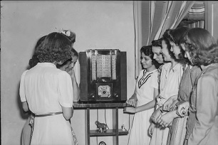 Foto em preto e branco de um grupo de jovens mulheres, em uma sala, em frente a um aparelho de rádio